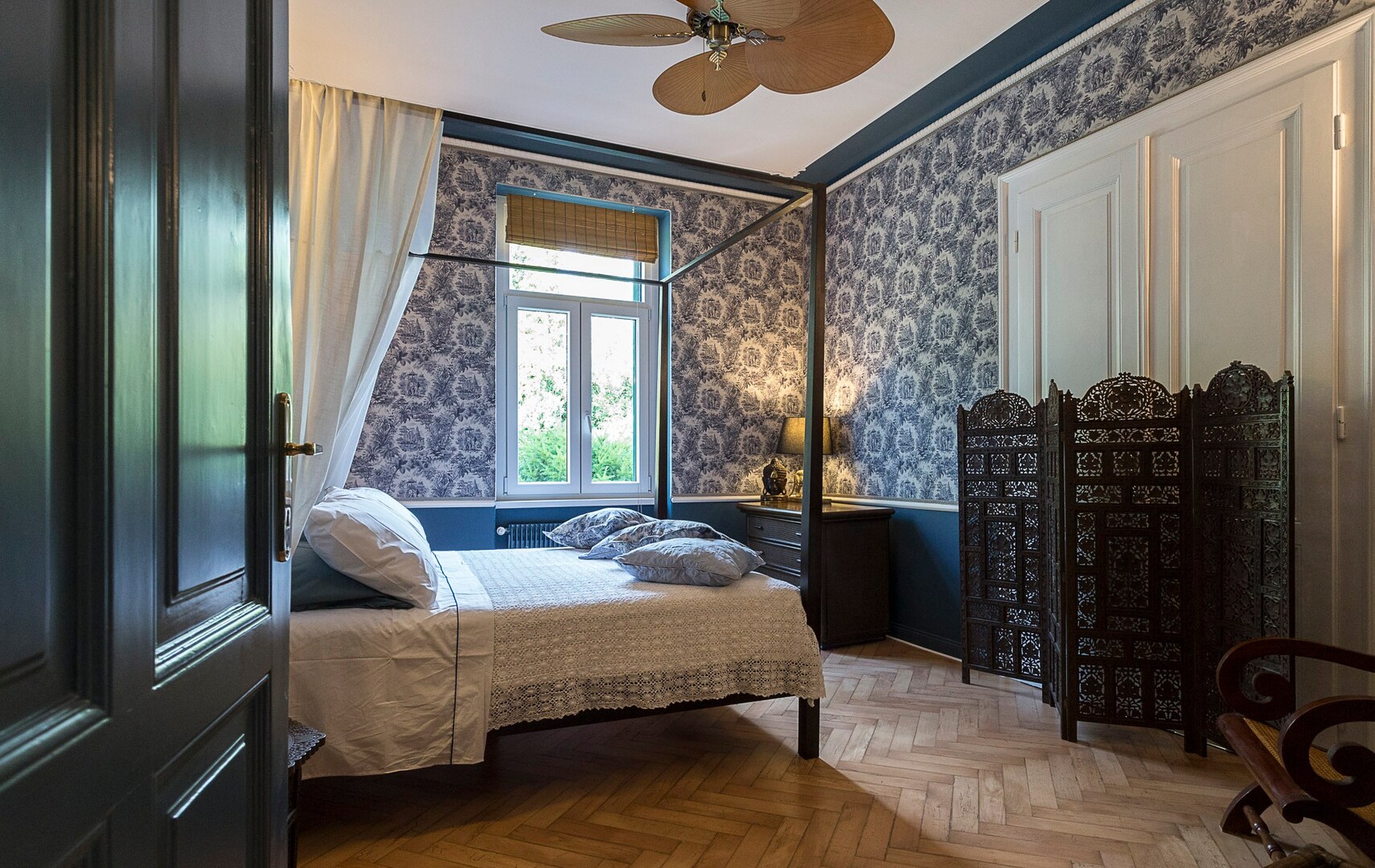 Bedroom with four-poster bed, patterned wallpaper, and wooden room divider.